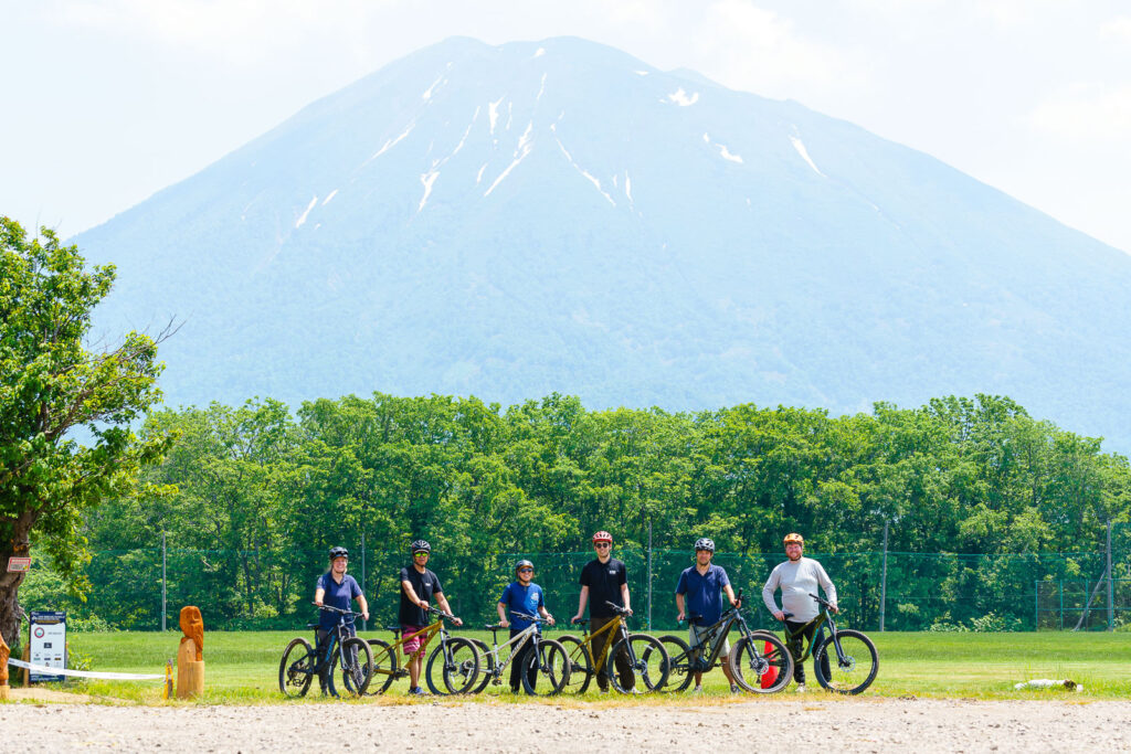 A group of six people with mountain bikes posing outdoors in front of a lush green forest and Mount Yotei in the background on a bright summer day.